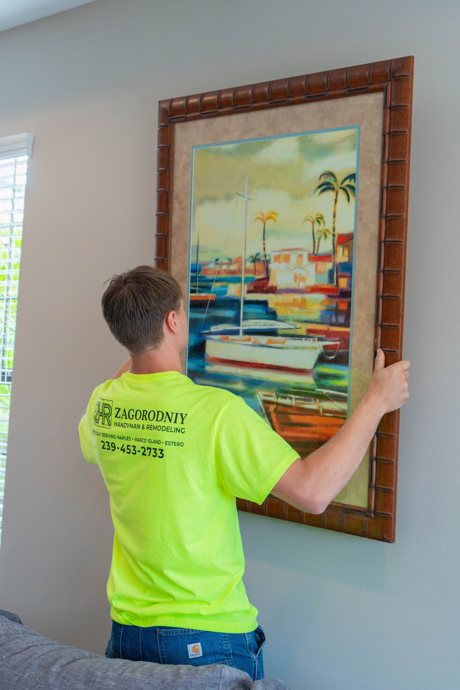 Handyman wearing a neon green shirt hanging a large picture on a grey wall in Naples.