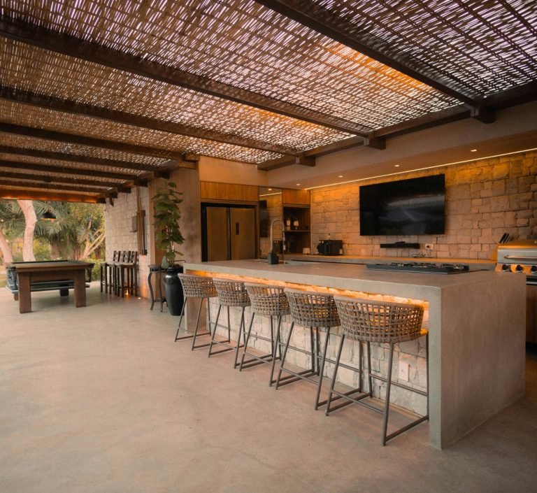 Outdoor kitchen and bar area with high stools, a countertop, and a television, under a shaded roof in Marco Island, FL.