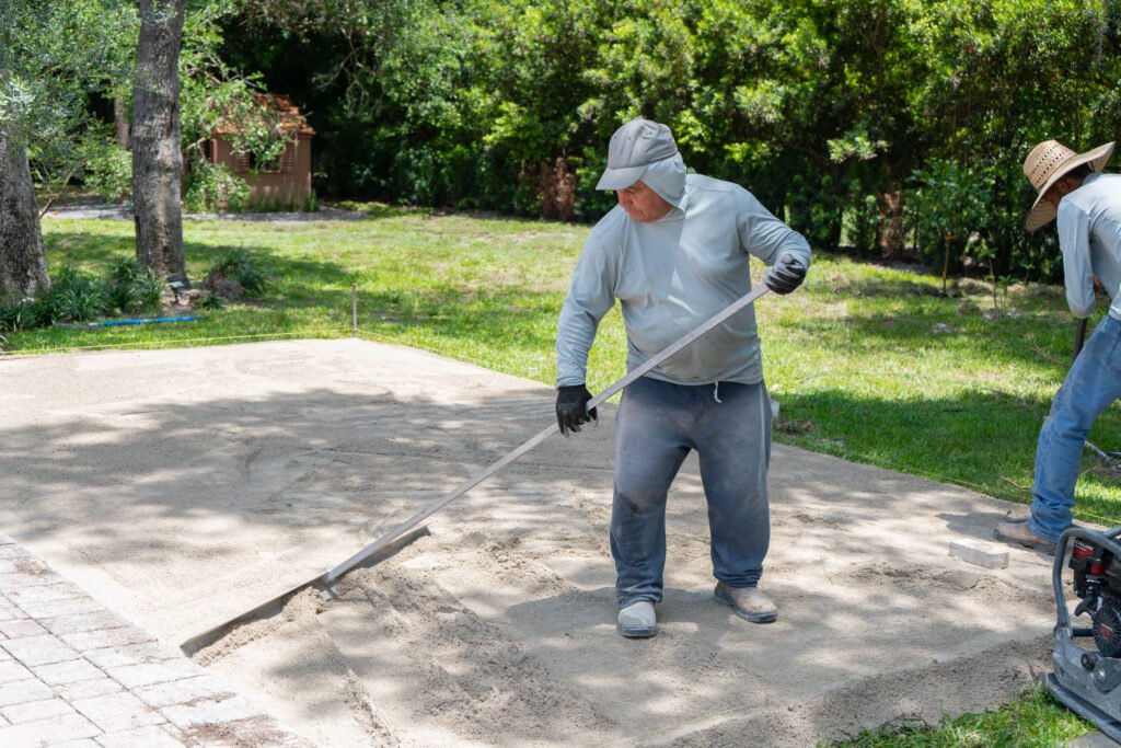 A worker using a long tool to level a pad of sand in a grey shirt and hat in North Naples.