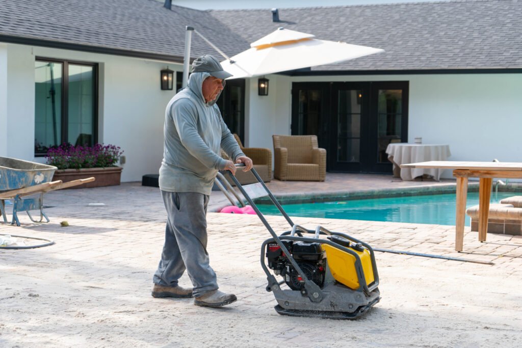 A worker in grey clothes using a machine to settle newly placed pavers with a pool behind him in North Naples.